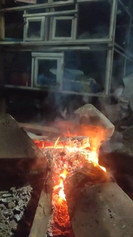 The video shows a wood-burning fire in a simple outdoor or semi-outdoor setting, likely at night or in low-light conditions. The fire is contained within some kind of makeshift hearth or fire pit, possibly surrounded by stones or bricks. A metal pot is placed over the fire, presumably for cooking. In the background, there's a wooden structure with compartments, possibly a storage unit or cage of some kind. There are no people visible in the video. The primary action is the fire burning and the implication of cooking or heating something in the pot.