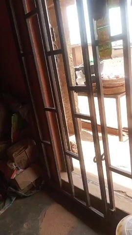 The video shows an interior view through a barred window. Outside, there's a small table with a woven basket on it, containing what looks like raw meat. Clothes or fabric are hanging on the bars of the window, partially obstructing the view. The outdoor area appears to be a dirt yard with some vegetation in the background, with glimpses of buildings in the distance. The lighting is bright outdoors, but the interior is dim. There are also household items visible in the lower corner of the interior.