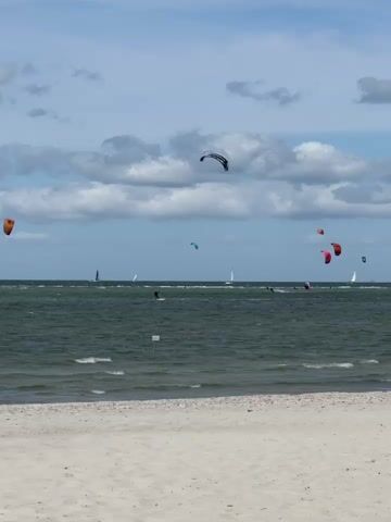 The video shows a beach with white sand in the foreground and a green ocean in the background. Several sailboats and kite surfers are visible on the water. The sky is blue with scattered white clouds. The kite surfers are in various positions, with some of them in the air. One kite surfer is using a black and white kite, another is using a red and orange kite, and another is using a red kite. There is a white signpost visible on the beach. The waves are small.