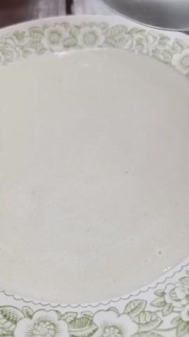 The video shows a close-up of a white creamy substance in a white bowl with a green floral pattern around the rim. A silver spoon is dipped into the substance, scoops some up, and then lifts it out, allowing the creamy substance to drip back into the bowl. The substance appears to be smooth and thick, with some small bubbles on the surface. The lighting is natural, suggesting an indoor setting.