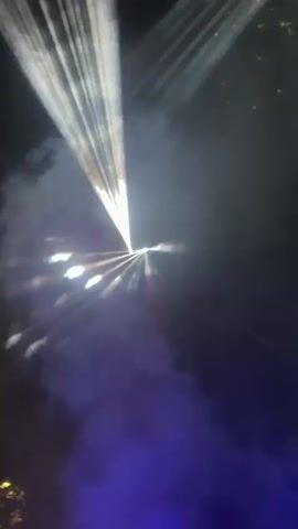 This is a short video taken from a low angle looking up at a dark sky filled with white and purple laser lights. The lasers are beams of light that fan out and create patterns in the smoky air, resembling a stylized figure eight or infinity symbol. Some of the beams are sharp and distinct, while others are diffused by smoke, creating a hazy and ethereal effect. The purple light appears to be coming from below, possibly from stage lighting. The scene is dark and atmospheric, with hints of foliage or structures at the very bottom of the frame. There are no people visible in the video.