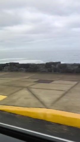 The video shows a roadside view of the ocean on a cloudy day. A yellow curb runs along the side of the road. A trailer with a metal cage is pulled by an unseen vehicle. A dark-colored pickup truck with a sign on its door is parked on the side of the road. The truck has a white sign on its door with text and numbers. The ocean is visible in the background with waves crashing against a stone wall. The road appears to be wet.