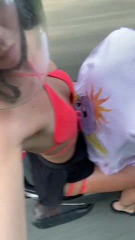 The video shows a woman riding a scooter, filmed from behind and above. She wears a red bikini top, black shorts, and a helmet with a clear visor. Her long, dark hair streams behind her in the wind. A white cloth with a sun design is draped over the back of the scooter. The road is gray asphalt and blurred with motion.