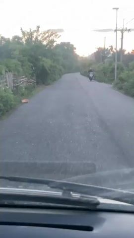 The video shows a view from inside a vehicle looking out through the windshield onto a paved road that curves to the left. On the right side of the road, a person is riding a white scooter in the distance. The road is bordered by lush green trees and foliage on both sides. The sky in the background is bright, suggesting it is either sunrise or sunset. There is a street light visible on the right side of the road. The windshield wipers are visible in the foreground.