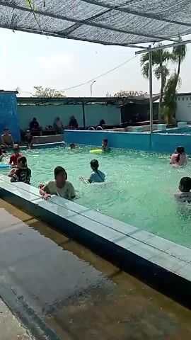 The video shows a group of children swimming in a pool on a sunny day. The pool is rectangular with blue tiles and a shallow end. There is a shaded area over a portion of the pool, supported by poles. Some adults are sitting and observing from the side of the pool. The water is clear and turquoise. The children are playing and splashing in the water.