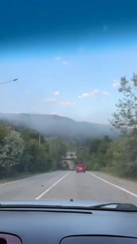 The video shows a view from inside a vehicle looking out through the windshield at a road. The road appears to be in a rural or mountainous area, with trees and vegetation on either side. In the distance, there is a red car driving on the road, and a stone bridge or archway. The sky is blue with some clouds and a hazy or foggy mountain range in the background. The sun appears to be low in the sky, possibly indicating early morning or late afternoon. The video is a single, continuous shot with no visible edits. The focus is on the road ahead and the scenery.
