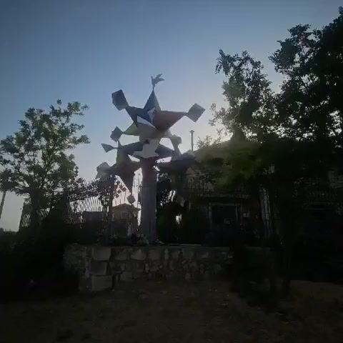 A multi-pointed, star-shaped metal sculpture, possibly a wind spinner or kinetic art piece, stands against a bright sky. The sculpture is supported by a pole, which is anchored to a low stone wall. The sculpture appears to be constructed from flat metal pieces, some of which are colored red, white, and blue. The scene is framed by trees on either side, with a building visible in the background. The lighting suggests it is either dawn or dusk.