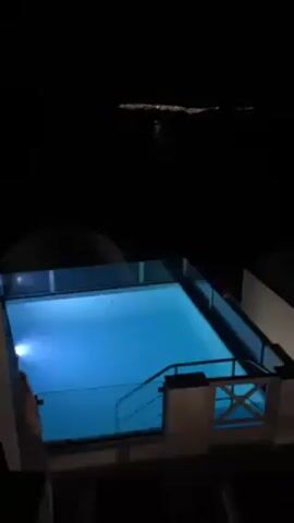 The video shows an outdoor view at night, likely from a balcony or elevated position. A small, blue-lit swimming pool is in the foreground. Behind the pool, there is a dark body of water and a distant coastline with lights. The camera pans to show a row of white buildings with distinctive architecture, possibly a village or town on a hillside. The overall environment appears calm and quiet. There are no visible people in the scene.