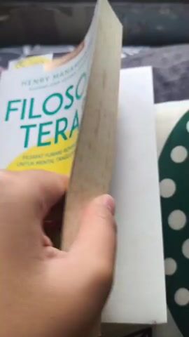The video shows a person's hand flipping through the pages of an open book. The book appears to be written in Indonesian. The left page is partially visible and seems to have a faded, textured appearance. The right page is more clearly visible and displays text in multiple columns, along with a small circular photograph of a smiling man wearing glasses and a light-colored shirt. The background of the scene is blurred, but suggests a surface with a striped pattern, possibly a blanket or bedding. The book's page numbers are visible, with one clearly showing "297" and another "205". The name "HENRY MANAMPIRING" is printed at the bottom of the right page.