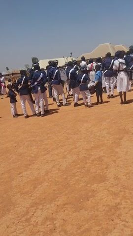 A band stands in formation on a dusty, reddish-brown field under a bright, clear sky. The band members wear matching uniforms of white pants and navy blue jackets with white trim. Many also wear white caps with navy blue trim. Some members carry musical instruments, primarily drums, including snare drums and a large bass drum. One band member is adjusting a snare drum that’s set on the ground. At least one person in a white dress is also part of the group. In the background, a large white tent is visible, along with other distant structures. The camera angle offers a view from behind the band, focusing on their backs and the surrounding open space.