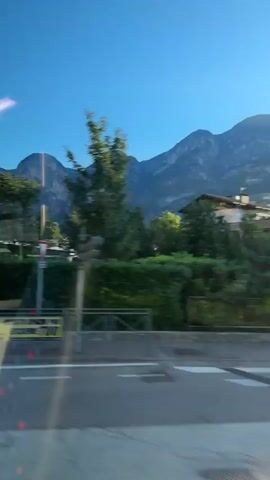 The video shows a fast-moving view from inside a vehicle, looking out at a passing landscape. The foreground is blurred due to motion, but several signs and a low hedge are visible. Beyond the hedge, a modern building with a distinctive yellow circular logo, likely a supermarket, is prominent. Trees, some of which are blurred with motion, are scattered around the building. In the background, large, rugged mountains dominate the scene under a clear blue sky. The lighting suggests it is daytime, possibly late afternoon or early morning, given the direction of the light and shadows. No people are clearly discernible due to the blur and distance, though the presence of a vehicle suggests human activity. The overall impression is of a journey through a semi-urban area with a backdrop of natural mountain scenery.