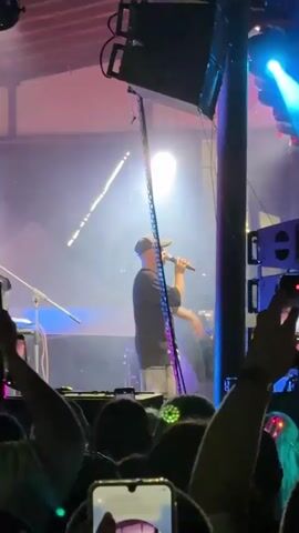 The video shows a man on a stage, holding a microphone and gesturing with his free hand. He is wearing a dark t-shirt, light-colored shorts, and a baseball cap. A bright spotlight shines on him from above, and there is smoke or haze in the air. Behind him, there are speakers and stage lighting. In the foreground, a crowd of people can be seen, many holding up their phones to record the performance. The atmosphere appears to be a live concert or event.