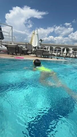 This media file captures a sunny day at an outdoor swimming pool, likely in Podgorica, Montenegro, given the context. The scene is dominated by the vibrant turquoise water of the pool, with a prominent water feature creating a cascading effect from a modern, curved metallic structure. In the foreground, a person wearing a bright neon yellow swimsuit swims through the water, their back to the camera.

The poolside is lined with lounge chairs and umbrellas, suggesting a resort or hotel setting. The weather appears clear and bright, with a blue sky filled with fluffy white clouds. The overall mood is one of leisure and enjoyment, typical of a vacation or a relaxing day by the pool. The sun is high in the sky, indicating it is likely midday. There is no visible text within the media file.