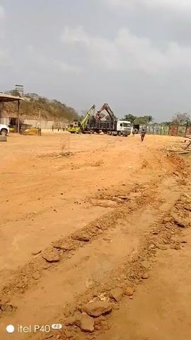The video shows a construction site with heavy machinery. In the center of the frame, an excavator is loading materials into a truck. A few people are visible near the machinery. The ground is mostly dirt and mud, with visible tire tracks. In the background, there is a structure with a roof, a white pickup truck parked under it, and what appears to be a hill. The sky is overcast. The camera slowly pans to the left. The “itel P40” watermark is visible at the bottom left corner of the screen.