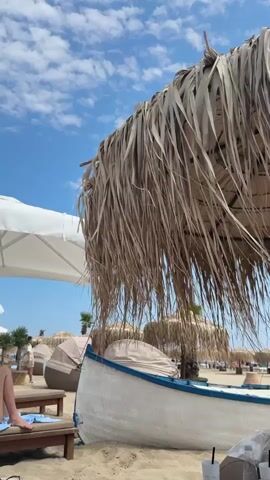 A person's legs are visible on a beach chair under a large straw umbrella. A white boat with a blue trim is on the sand next to the umbrella. In the background, there are other beach chairs, umbrellas, and a few people walking. The sky is blue with some clouds.