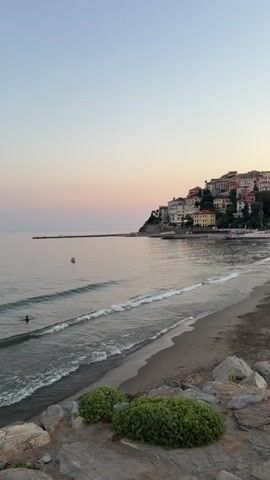 This vertical video captures a serene coastal scene at dusk in Imperia, Italy. The sky transitions from a pale blue overhead to soft hues of pink and lavender near the horizon, indicating the late evening hour. A calm sea laps gently onto a sandy beach, with small waves breaking near the shore. 

In the water, two figures can be seen swimming or wading, enjoying the tranquil waters. Further out, another person appears to be standing in the water. A long pier extends into the sea on the left side of the frame, and beyond it, the coastline is dotted with colorful buildings clustered on a hillside, characteristic of Italian coastal towns. Lush green bushes and large rocks frame the foreground of the beach.

The overall mood is peaceful and idyllic, with the soft light of sunset creating a warm and inviting atmosphere. There is no visible text in the video.