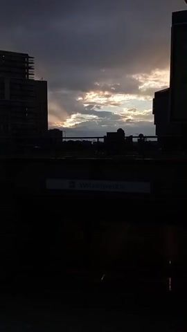 The video shows an exterior view of a subway station entrance in an urban setting. The station is labeled "Wilanowska". The sky above is cloudy with some sunlight breaking through, creating sunbeams. There are buildings visible in the background, including modern high-rise structures and some older buildings. The entrance to the subway station consists of a set of stairs leading down into a tunnel. A sign with the station name "Wilanowska" is posted above the entrance. There is also a large billboard advertising Tymbark. The overall scene appears to be captured during either sunrise or sunset, given the lighting conditions.