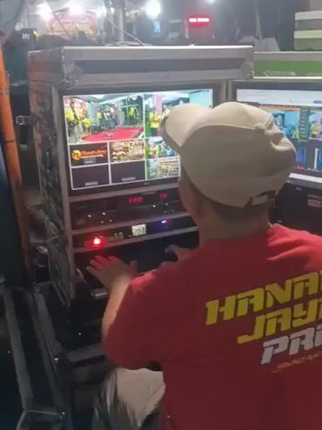 The video shows a person wearing a red shirt with yellow lettering that reads "HANAM JAYA PRO SOUND RACING" and a light-colored baseball cap, facing away from the camera and operating a complex audio-visual setup. The setup consists of multiple racks filled with electronic equipment, including amplifiers and processors with illuminated indicator lights. A large monitor displays multiple video feeds, seemingly showing different camera angles of an event. To the right, another monitor shows what appears to be a control interface with various input and output channels. The environment appears to be outdoors at night, with some lighting from the equipment and possibly overhead tents or structures visible in the background. Other people are faintly visible in the background, suggesting this is a live event. The person is actively engaged with the controls, their hands near a keyboard or console.
