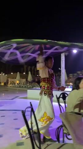 Here is a description of the video:

The video shows a man performing a traditional Egyptian Sufi dance, known as Tanoura, at night. He is wearing a white robe-like outfit with colorful embroidery. He spins rapidly while holding a large, round, illuminated skirt that billows out around him. 

The performance takes place outdoors, likely near a pool area at a resort. People are seated at tables nearby, watching the performance. One woman is seen assisting a little girl, encouraging her to stand up and watch. The area is lit with blue and purple lights, enhancing the atmosphere.
