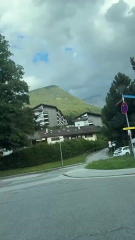 The video is a first-person perspective from a vehicle moving through a scenic mountainous region. The first shot shows a traffic sign with an arrow indicating a turn to the right, a lamppost with some overgrown grass around its base, and a street. The second shot shows a billboard advertising a Christmas market event with dates. The third shot is of a house with a well-maintained garden and a mountain in the background. The fourth shot shows trees and a stone wall along the side of the road. The sky is partly cloudy throughout the video. The overall impression is a drive through a picturesque rural or small-town European setting.