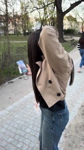 The video shows a park-like setting with a cobblestone path. A woman wearing a tan jacket and jeans walks along the path.  The camera pans, showing people sitting on benches, trees, and buildings in the background. A trash can is visible near the path. The sky is partially cloudy.