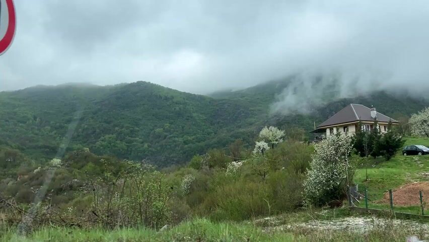 This video captures a panoramic view of a lush, mountainous landscape in Obzovica, Montenegro, likely taken from a moving vehicle. The foreground features a silver guardrail, indicating the perspective from a road or overlook.

The dominant subjects are the verdant mountains, thickly covered in trees and vegetation, predominantly shades of green, with some lighter green foliage suggesting new growth or different species. White blossoms are scattered among the trees, indicating spring or early summer.

The mountains recede into the distance, with the upper slopes shrouded in thick, low-hanging clouds and mist, giving the scene a soft, ethereal quality. The weather appears overcast and cloudy.

On a hillside to the right, a two-story house with a brown roof and light-colored walls is nestled into the terrain, surrounded by trees. Another smaller house is visible further down in the valley to the left of the main house.

The activity involves a slow, panning movement of the camera, capturing the expansive natural beauty of the region. No people are visible. The overall mood is tranquil and serene, despite the cloudy conditions. There is no visible text in the video.