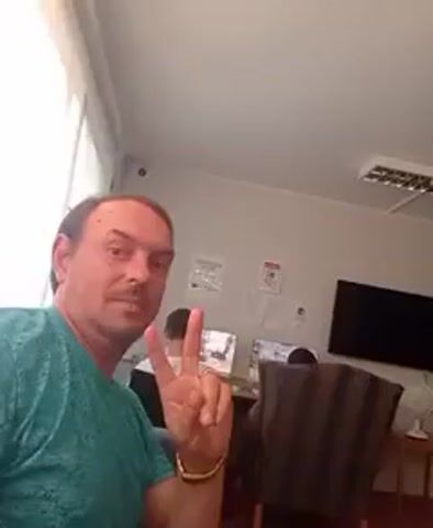 The video shows a man in a teal shirt taking a selfie in an indoor space. He is making a peace sign with his right hand. In the background, two individuals are seated at computers, and there is a television mounted on the wall. The room appears to be a common area or lounge with chairs and a table. The lighting suggests it is daytime.