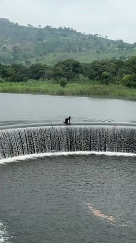 A person is standing on top of a concrete dam with a waterfall cascading down one side. The person is facing away from the camera and appears to be holding their hands up. The dam is surrounded by lush green vegetation and a hill in the background. The sky is overcast.