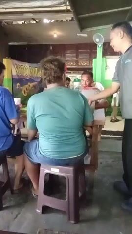 The video shows a group of people in what appears to be an outdoor eating area. In the foreground, a person wearing a teal shirt and denim shorts is seated on a brown plastic stool, facing away from the camera. To their left, another person in a blue t-shirt and dark shorts is also seated on a similar stool. In the background, a man in a gray shirt is standing and leaning over a table, interacting with someone seated at the table. Another person, possibly a child, is visible in the far background. A banner with yellow sunflowers and the text "SOTO ROU" is hanging on the wall behind the seated individuals. The environment appears rustic, with exposed beams overhead and concrete flooring.