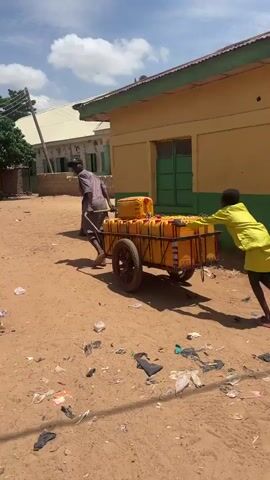 Two people are pushing a cart loaded with yellow containers down a dirt road. The person in front is wearing a brown shirt and pants and is pulling the cart by its handles. The person behind is wearing a bright yellow shirt and shorts and is pushing the cart from behind. The cart has two wheels and is loaded with at least ten yellow containers, which appear to be large jerrycans. The road is sandy and littered with debris. In the background, there are buildings with yellow walls and green trim, as well as trees and power lines. The sky is blue with scattered clouds.