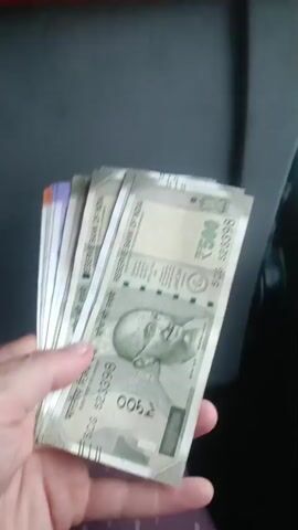 A hand is holding a fan of Indian Rupees banknotes. The majority of the visible banknotes are 500 rupee notes, featuring a portrait of Mahatma Gandhi. The hand is holding the banknotes in a fanned out position, and it appears to be moving slightly. The background is dark and indistinct, suggesting an indoor or dimly lit environment. The person holding the money is wearing a purple and white polka dot garment. The banknotes appear to be new and in good condition. The lighting is dim.
