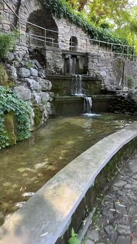 A cascading waterfall tumbles over multiple stone levels into a shallow, murky pond. The pond is bordered by a curved, concrete edge in the foreground, with a stone pathway visible to the right. The waterfall structure itself is built into a stone wall adorned with greenery. In the background, a stone archway and railing suggest a pathway or terrace. Sunlight filters through the trees above, casting dappled light on the scene. The water appears slightly green and contains some debris. No people are visible in the video.