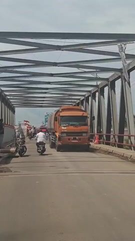 A long shot captures a bridge with metal trusses overhead. A large orange tanker truck is the prominent object, partially obscuring the view of the road. In front of the truck, several motorcycles and a red car are visible. A person is riding on the back of one of the motorcycles, and other motorcycles are in motion. The road surface appears to be asphalt. The weather seems overcast, with a bright sky suggesting daylight.