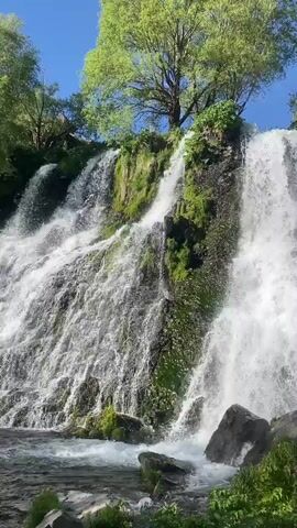 The video shows a multi-tiered waterfall cascading over rocks covered with moss and vegetation. The sky is a clear blue, and trees are visible above and around the waterfall. A person briefly appears with arms outstretched in front of the waterfall. The video then pans upwards towards the sun, briefly showing other people in the area. The setting appears to be a natural, outdoor environment.
