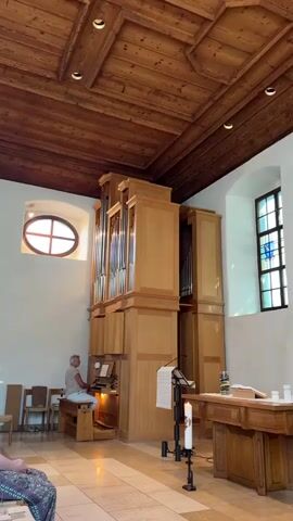 A man sits at an organ in a church and plays it. The organ is large and made of wood. The church has a wooden ceiling and a tiled floor. There are chairs and a table with candles in the church. A stained glass window is visible on the right side of the church.