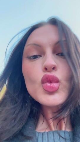 A woman with long dark hair is outdoors. She is wearing a grey sweater and jacket. She is close to the camera. First, she is making a pucker face and looking upwards. Then, she looks from side to side. Finally, she smiles at the camera, showing her teeth. Her hair blows in the wind. Trees and a clear blue sky are visible in the background.