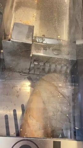 A person's hand inserts a loaf of bread into a commercial bread slicer. The bread is placed in the slicing mechanism, and the machine begins to slice it. Crumbs are visible on the machine and in the surrounding area.