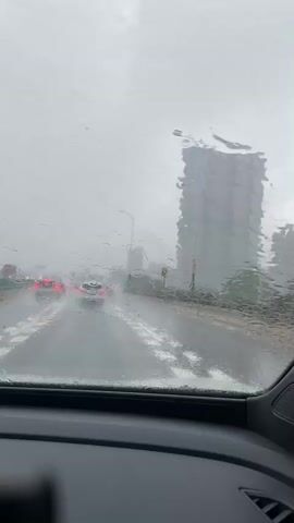 The video shows a view from inside a car on a rainy day. Raindrops are visible on the windshield, and the wipers are intermittently active, clearing the view. Multiple cars are driving on a wet highway, with their brake lights illuminated. Water is splashing on the road due to the rain and the movement of vehicles. In the background, a tall, modern building is partially obscured by the rain and fog. The overall environment appears to be a city highway during a heavy downpour.