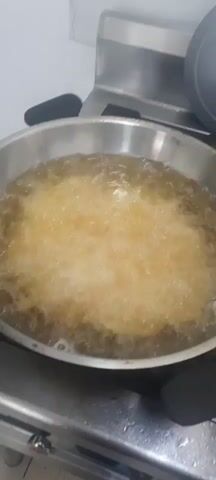 The video shows a close-up of a pan of oil with food being fried. The oil is bubbling vigorously. To the right of the pan, there is a metal bowl filled with what appears to be french fries. Behind the pan and bowl, there are two large pots. One pot contains a reddish-orange stew or soup. The other pot is partially visible and appears to be made of stainless steel. The overall environment looks like a kitchen, possibly a commercial one, given the stainless steel surfaces and equipment. There are no people visible in the video. The lighting is bright and consistent with kitchen lighting.