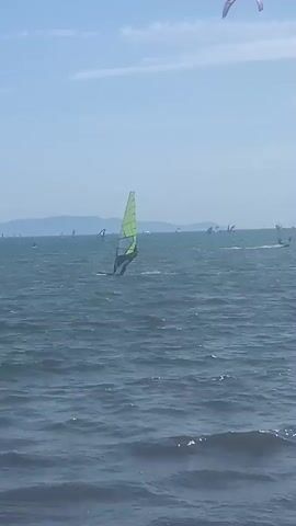 A wide shot shows a choppy, dark blue ocean under a clear blue sky with wispy clouds. In the foreground, ripples and small waves are visible. In the middle ground, a lone person is windsurfing, with a bright yellow-green sail on a dark board. The person is positioned in profile, facing left. Further out in the water, numerous other windsurfers and kitesurfers are scattered across the horizon. Some kites are visible in the sky, including a large, colorful kite with a curved shape on the far right and a smaller, white kite to the left of it. In the far distance, a hazy, blue mountain range forms the horizon. The overall scene depicts a windy day at sea, with various water sports activities taking place.