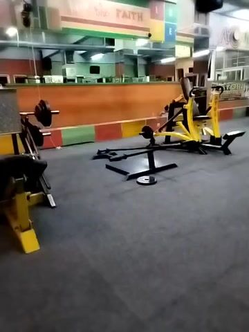 The video shows an empty gym with various exercise equipment. In the foreground, there is a yellow weight machine and a barbell with weights. Further back, there is a punching bag hanging from the ceiling and other weight machines, including what appears to be a cable machine. The gym has a grey carpeted floor and fluorescent lighting. The walls are decorated with colorful stripes and text that reads "FAITH" and "HOPE." There are also what appear to be pool tables in the background, suggesting it might be a multi-purpose facility. The gym appears to be clean and well-maintained, but unoccupied.