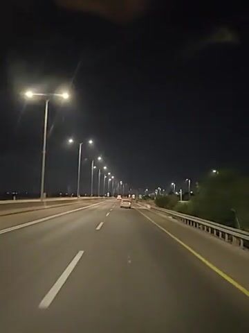 The video shows a nighttime view from inside a vehicle traveling on a highway. Streetlights illuminate the road, which has multiple lanes and a yellow dividing line. There is one other car visible ahead in the distance. The sky is dark, indicating nighttime. The environment appears to be a developed area with some vegetation on the right side of the road.
