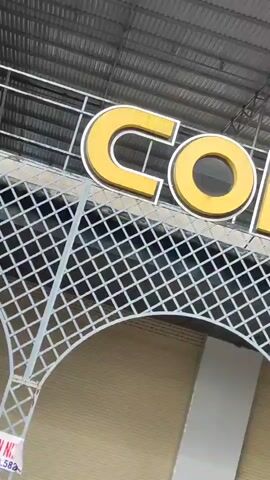 The video shows a close-up of a large yellow sign that reads "COFEE" in bold, uppercase letters. The sign is illuminated from behind and appears to be mounted above an entrance to a building. Below the sign is a white, ornamental lattice archway. The background consists of a corrugated metal roof and the exterior wall of the building, which is a light tan color. The lighting suggests it is daytime, and the camera angle is looking upwards at the sign. The footage is shaky, indicating it was likely filmed handheld.