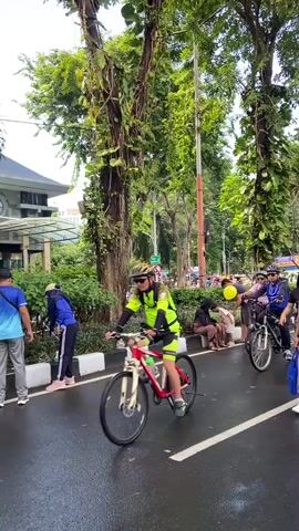 The video shows a street scene with a crowd of people on a bicycle route. The crowd includes people walking and riding bicycles. Some are riding regular bicycles, while others are riding modified bikes. The crowd contains people of all ages. The street is wet. Tall trees line the street. There is a building partially visible in the background.