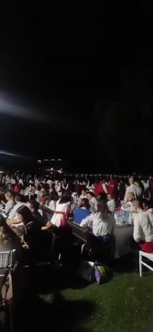 A night-time outdoor gathering is depicted with many people seated at long tables, eating and socializing. The tables are set with plates, glasses, and bottles, and some people are wearing traditional clothing in red and white. The scene is illuminated by bright lights and string lights, creating a festive atmosphere. The background is dark, with some distant lights suggesting buildings or structures. The camera pans slightly, revealing the scale of the event with numerous attendees spread across the area.