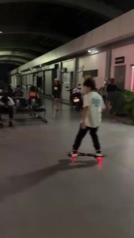 A young person on a skateboard with illuminated wheels rides in a dimly lit covered area, possibly a bus terminal. The skateboarder wears a white t-shirt with a graphic and dark pants. In the background, several buses are parked, and other people are visible, some of whom are blurred due to motion. The skateboard's wheels emit a red glow as it moves across the polished floor.