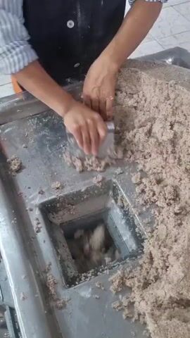 This is a video clip that shows a close-up of a person's hands working with what appears to be a meat processing machine. The machine has a large, stainless steel surface with a rectangular opening where a mixture of ground meat is being fed. A metal scraper is being used to push the mixture into the opening. In the background, there is a large metal bowl filled with what looks like raw sausages, submerged in water. The setting appears to be a commercial kitchen or food processing facility, with tiled floors and stainless steel equipment. The lighting suggests it could be daytime. There are no visible signs of weather conditions. The activity clearly depicts food preparation, specifically the process of making sausages. The focus on the hands and the machinery suggests a deliberate demonstration or a worker engaged in their task. There is no discernible text within the video itself.