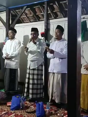 The video shows an indoor gathering. Three men are standing and facing forward. They are all wearing traditional Islamic attire, including white shirts and patterned sarongs. The man in the center is holding a microphone and speaking, while the other two men are holding books. They are standing on a patterned rug, with blue bags and food items arranged on the floor in front of them. The background shows wooden walls and a ceiling with exposed beams. Other people are visible in the background, some of whom are also wearing traditional clothing. The lighting is dim, creating a warm and intimate atmosphere.