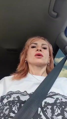 The video shows a woman with light pink hair and bright red nail polish, wearing a white t-shirt with a black lace design. She is inside a car, with a seatbelt across her chest. The woman appears to be talking, as evidenced by her mouth movements and hand gestures. She makes a peace sign with two fingers and points towards the camera at different points in the video. The lighting suggests it is daytime, and the reflection in the car window shows a cloudy sky, hinting at overcast weather. The scene is likely a typical roadside setting, with trees visible in the background outside the car window. The woman's facial expressions could be interpreted as conveying a range of emotions, from thoughtful to playful. No visible text is present in the video.