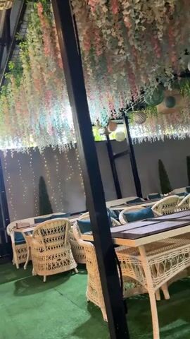This is a video file capturing an outdoor seating area, likely a cafe or restaurant patio, adorned with decorative elements. The scene is set under a covered roof structure, with tables and chairs made of woven material, possibly wicker, arranged on a green, artificial turf floor. The tables are set with placemats, and some chairs have cushions.

The area is decorated with hanging floral arrangements in shades of pink, white, and green, creating a whimsical and romantic ambiance. String lights are draped along a back wall, adding a festive glow, especially as the lighting suggests it might be evening or an indoor setting that mimics evening. Several paper lanterns of various sizes and colors are suspended from the ceiling, further enhancing the decorative theme. Two small, conical evergreen trees are placed strategically in the background.

There are no people visible in the video, and no specific activity or event is taking place; it appears to be a static shot of the environment. The overall mood conveyed is tranquil and inviting, with a focus on aesthetic appeal and creating a pleasant atmosphere for diners. There is no visible text in the video. Given the location context provided as Karaganda, Kazakhstan, this scene likely depicts an outdoor dining space within that city.